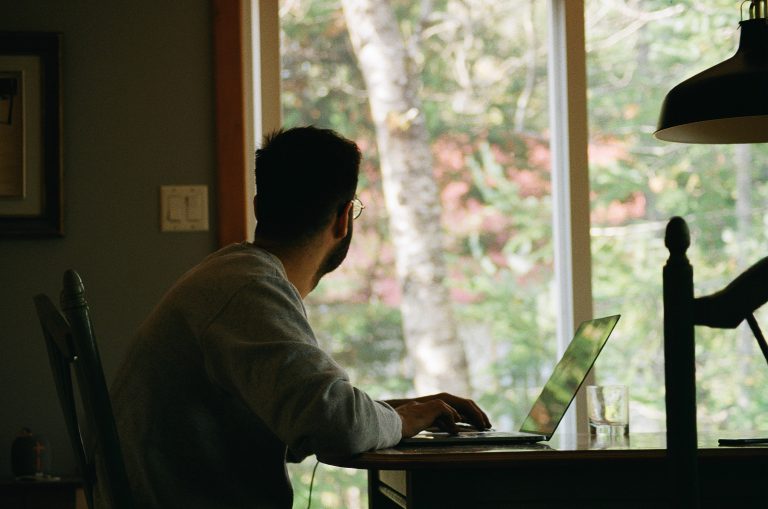 A man stares out of a window while sitting at a laptop